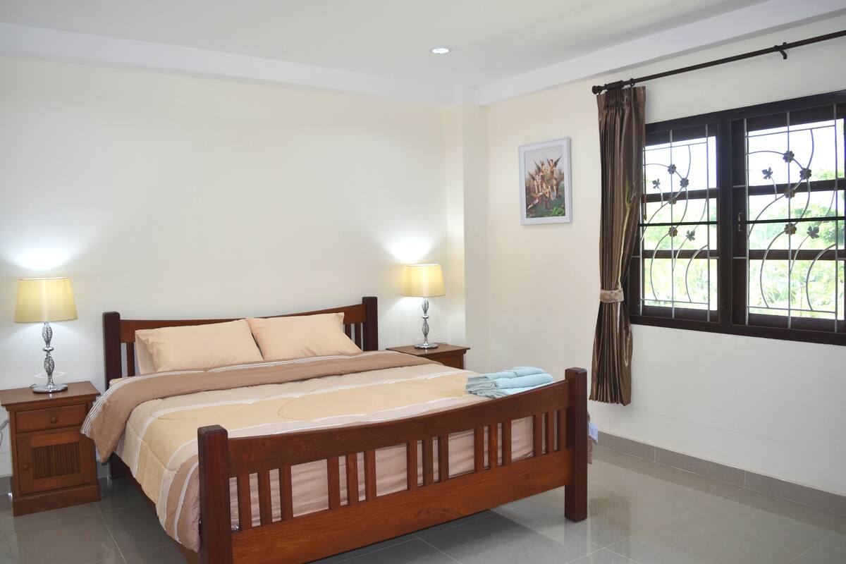 Quarto arrumado com cama de madeira, duas mesas de cabeceira com abajures, uma janela com cortinas e um quadro na parede.
