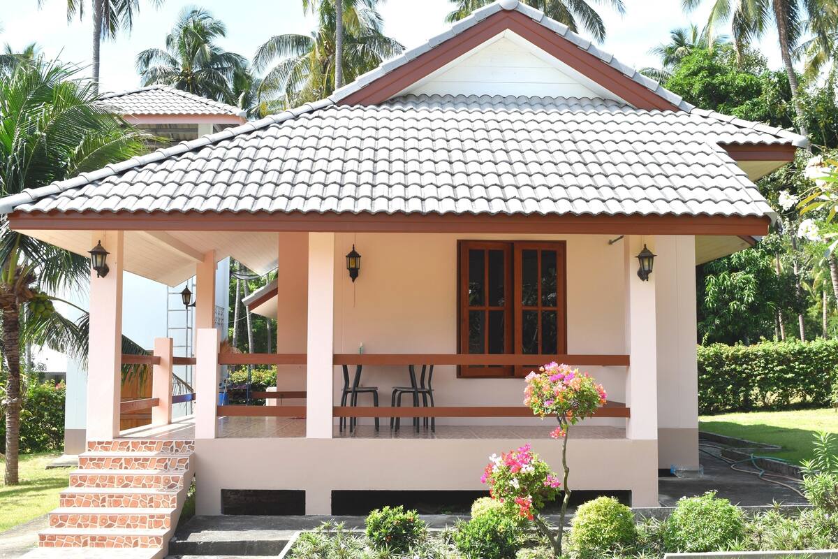 Uma casa de dois andares com telhado em telhas, varanda com cadeiras e um pequeno jardim com flores.
