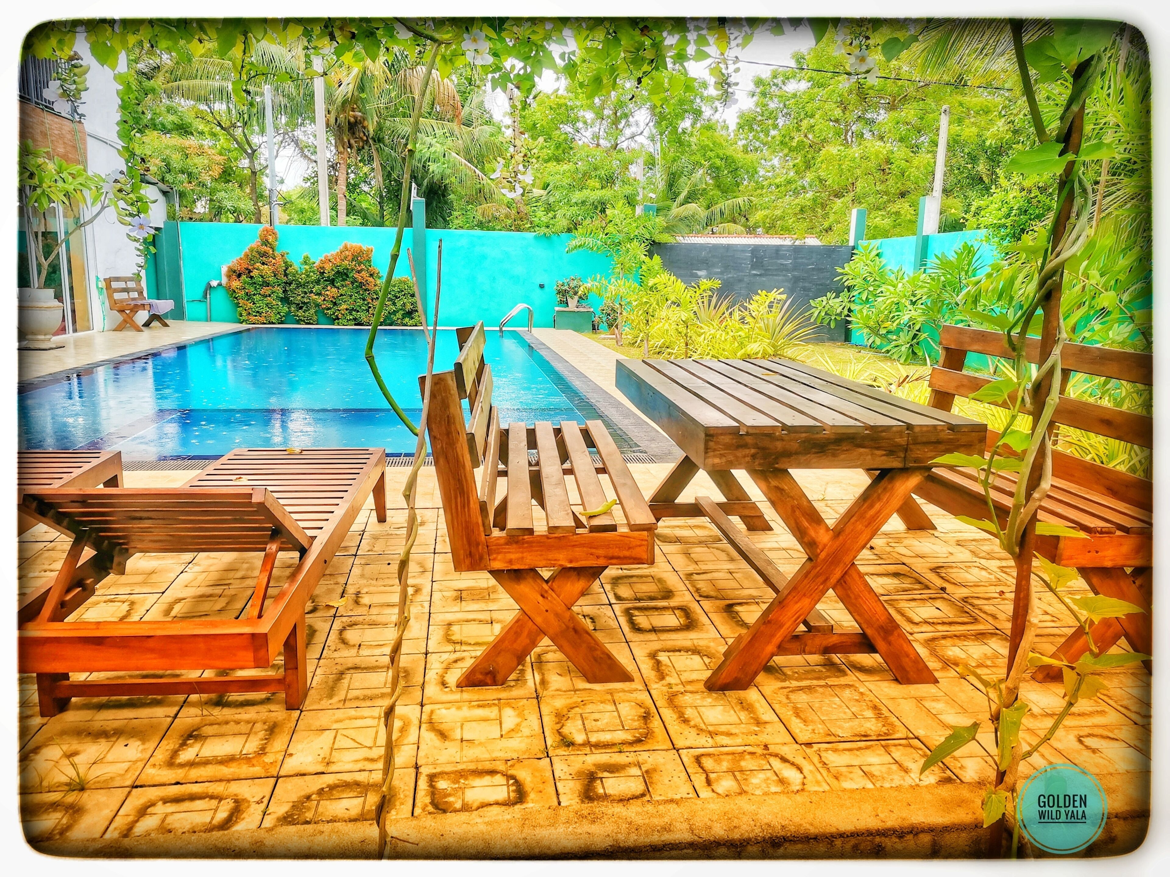 Zona de piscina con tumbonas de madera y una mesa, rodeada de exuberante vegetación y un muro azul.