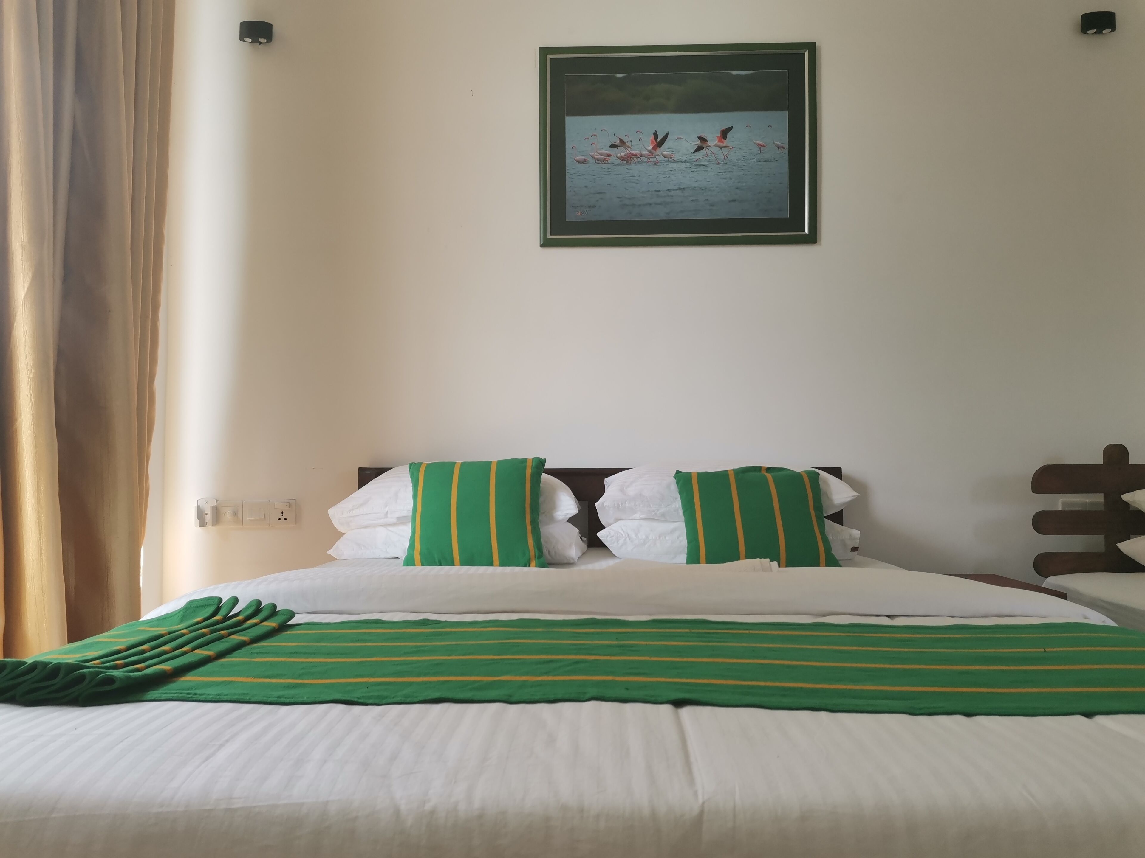 Una cama bien hecha con ropa de cama verde y blanca, un cuadro enmarcado de flamencos en la pared y un enchufe en la pared.