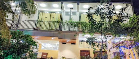 A multi-story building with balconies, a well-lit entrance, and a fenced garden area.