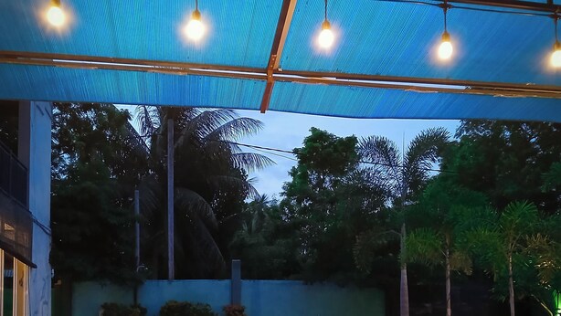 An outdoor swimming pool with a blue-lit water feature, surrounded by lounge chairs and a covered area with hanging lights.