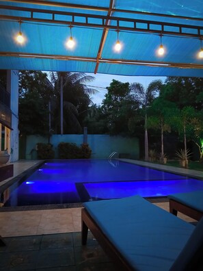 Een buitenzwembad met een verlicht waterkunstwerk in blauw, omringd door ligbedden en een overdekt terras met hangende lampen.