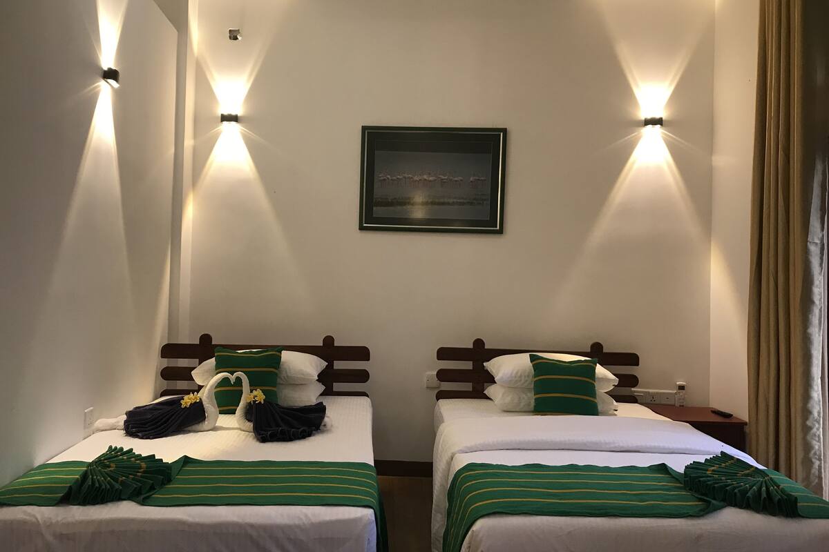 Twee eenpersoonsbedden met groen-wit beddengoed, een ingelijst schilderij aan de muur en wandlampen.