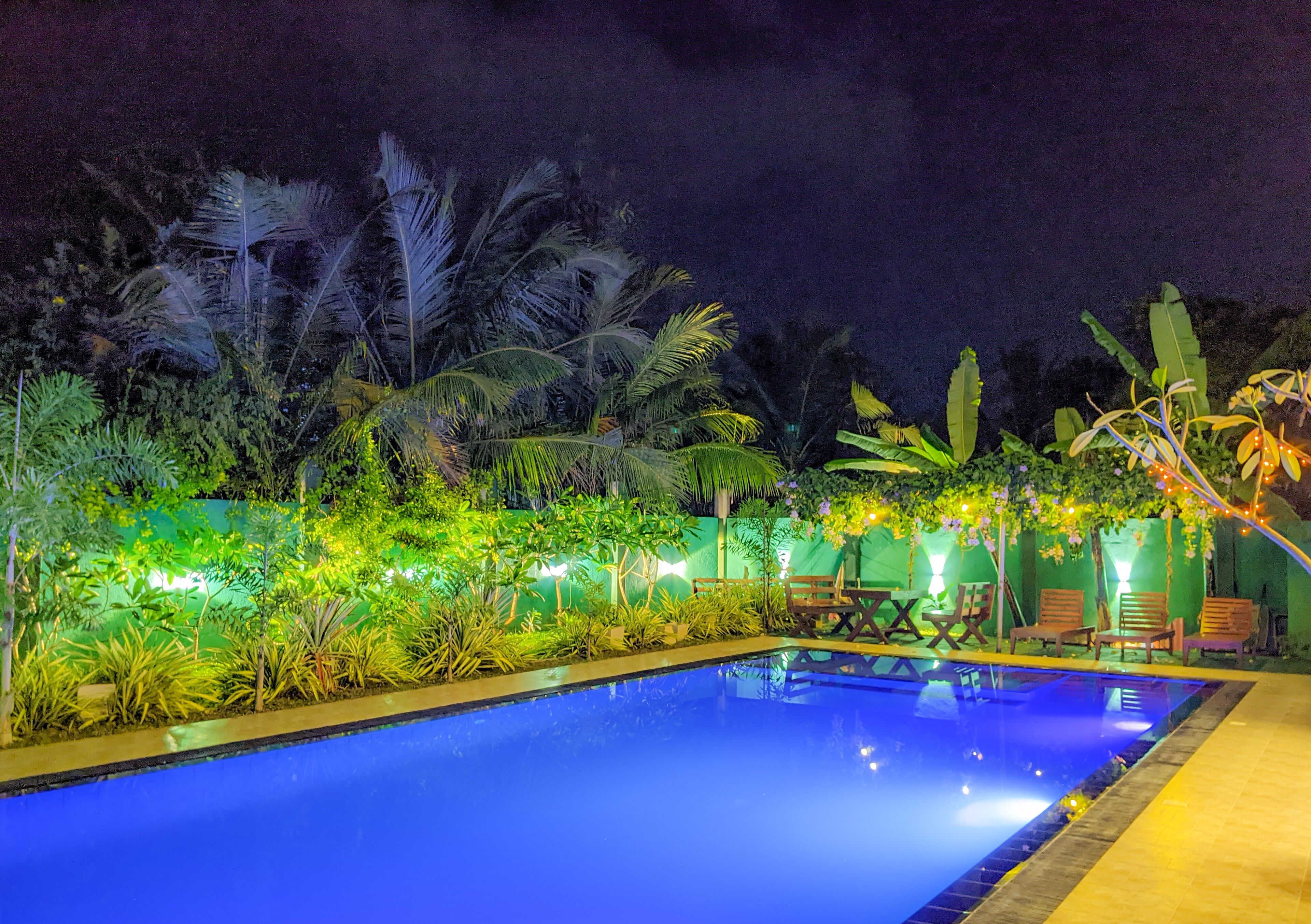 Une piscine nocturne entourée de verdure, avec des espaces aménagés pour s’asseoir.