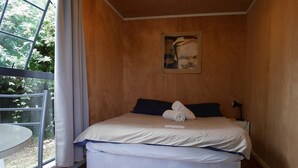 Una cama individual con colchón blanco y ropa de cama azul, una mesita de noche pequeña con lámpara, y una ventana con cortinas.