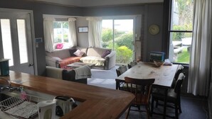 Una sala de estar acogedora con sofá, una mesa de comedor con sillas, una zona de cocina con fregadero y hervidor, y un amplio ventanal con cortinas.