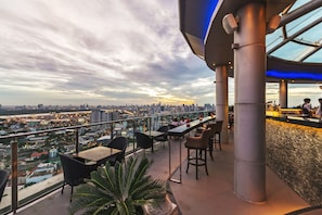 A rooftop bar with tables and chairs, overlooking a cityscape with buildings and a sunset.