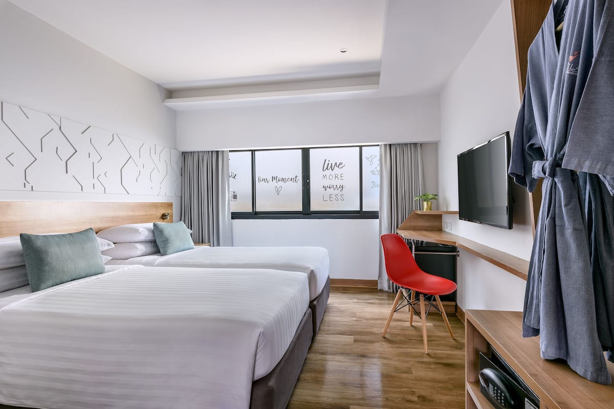 Quarto de hotel moderno com duas camas, televisão, secretária e uma cadeira vermelha.