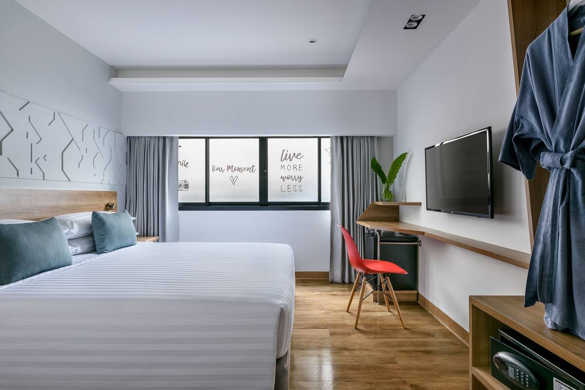 Quarto de hotel moderno com uma cama grande, televisão, escrivaninha e janela com frases motivacionais.