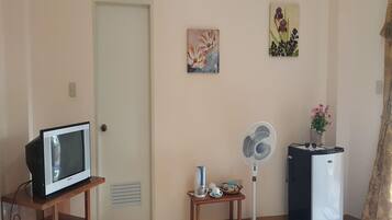 Una sala de estar con un televisor sobre un soporte de madera, un ventilador de pie, un refrigerador, una mesita con un jarrón de flores y dos cuadros colgados en la pared.