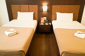 Habitación de hotel con dos camas, cada una con una almohada y una toalla doblada. Entre las camas hay una mesita de noche con una lámpara, un teléfono y un libro.