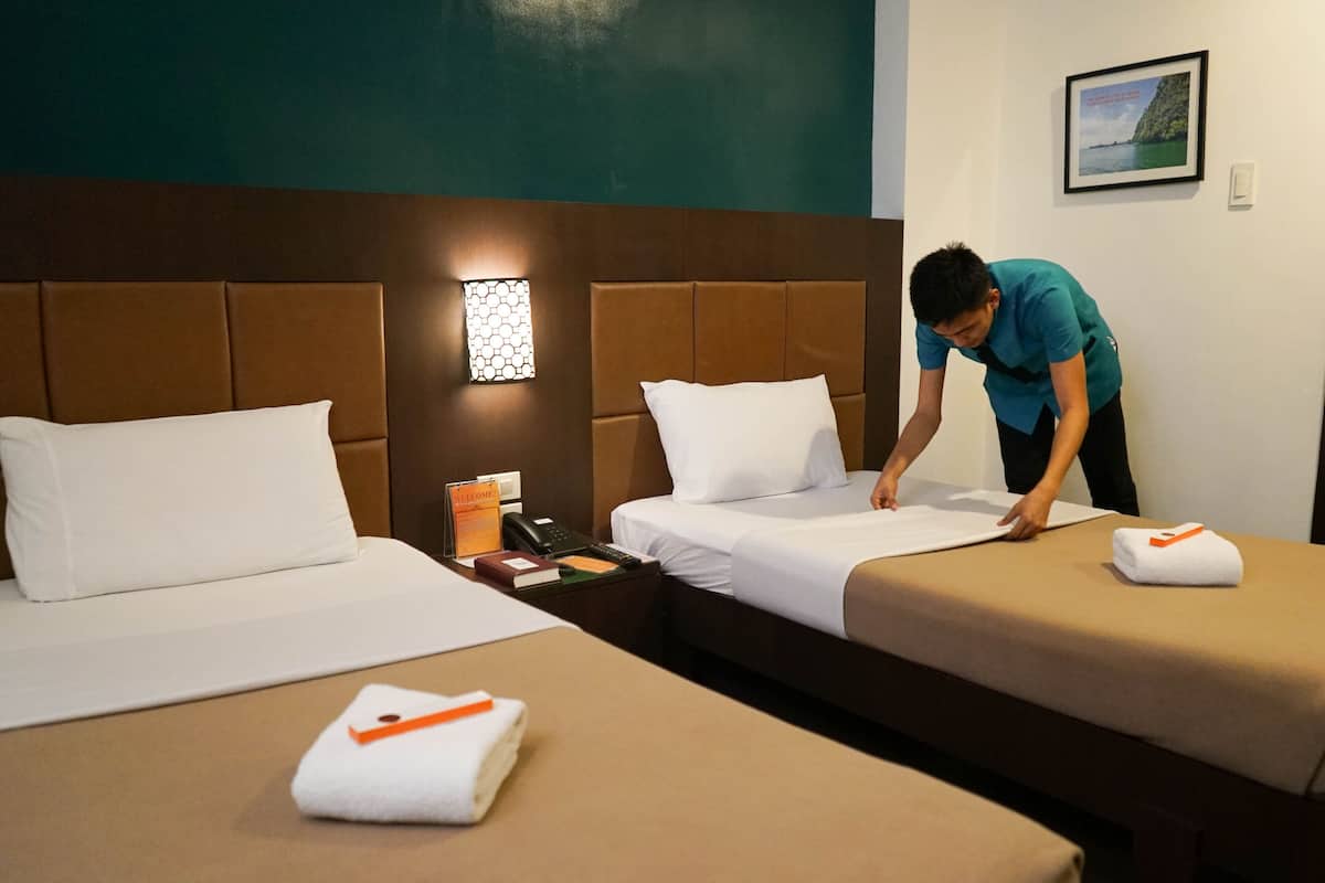 Una habitación de hotel con dos camas, una mesita de noche con un teléfono y una caja de bienvenida, y una lámpara fijada a la pared.