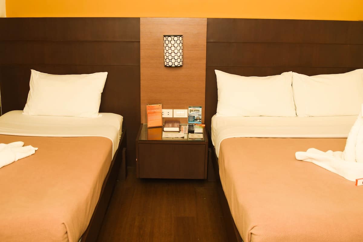 Habitación de hotel con dos camas, cada una con un almohadón blanco y una toalla con forma de cisne. Hay una mesita de noche entre las camas con un cartel de bienvenida y una pequeña mesa con un libro y una caja.
