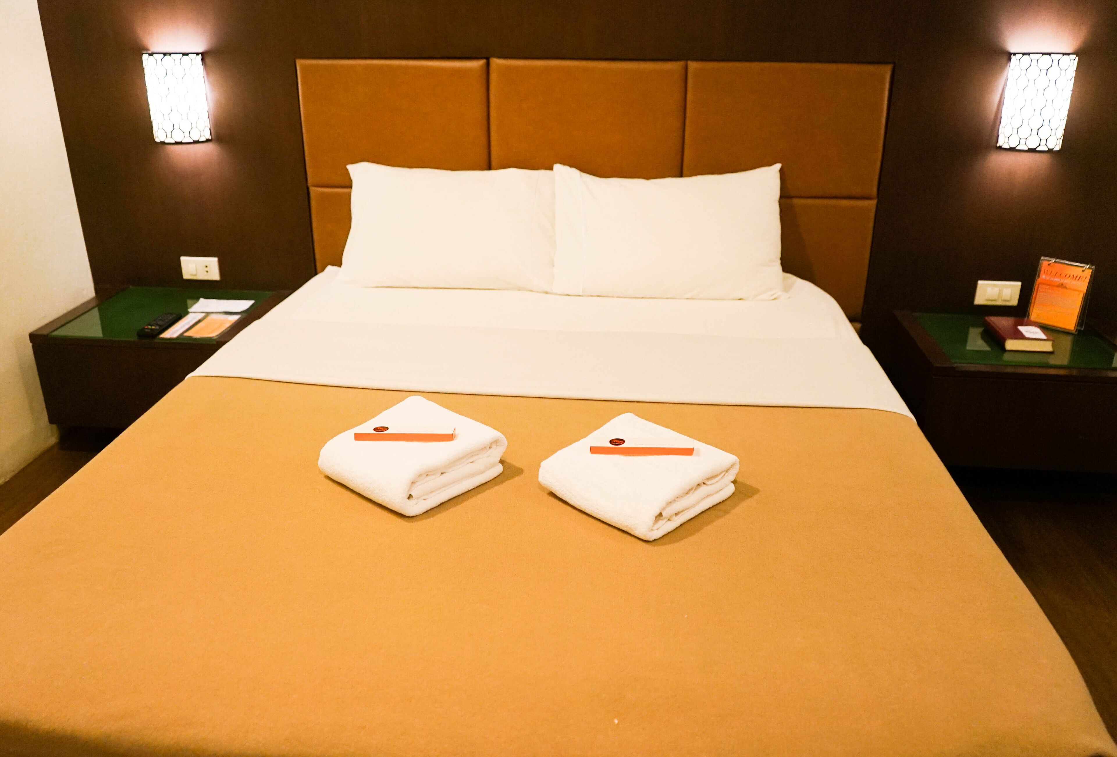 A hotel room with a large bed, two pillows, a brown headboard, two nightstands, and two wall-mounted lamps.
