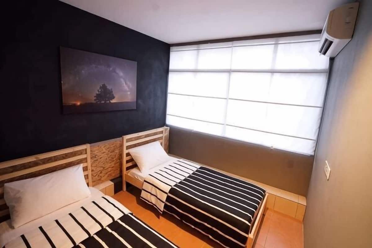 Ein Schlafzimmer mit zwei Betten, einem Fenster mit Jalousien, einem Bild an der Wand und einer Decken-Klimaanlage.