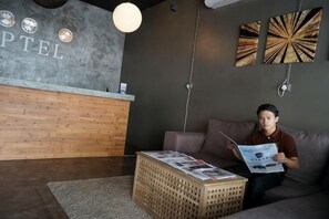A reception area with a wooden counter, a guest sitting on a couch reading a magazine, and a decorative wall with abstract art. - Koptel Budget Hotel (Kuching)