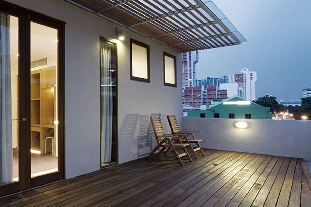 Un balcón moderno con piso de madera, una puerta de vidrio que da a una habitación interior y una vista panorámica de la ciudad al fondo.