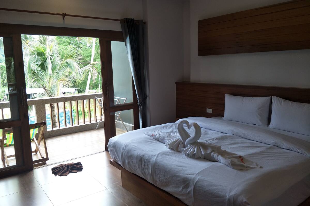 Une chambre d’hôtel avec un lit, une tête de lit en bois, un balcon donnant sur des palmiers, et une paire de serviettes blanches disposées sur le lit.
