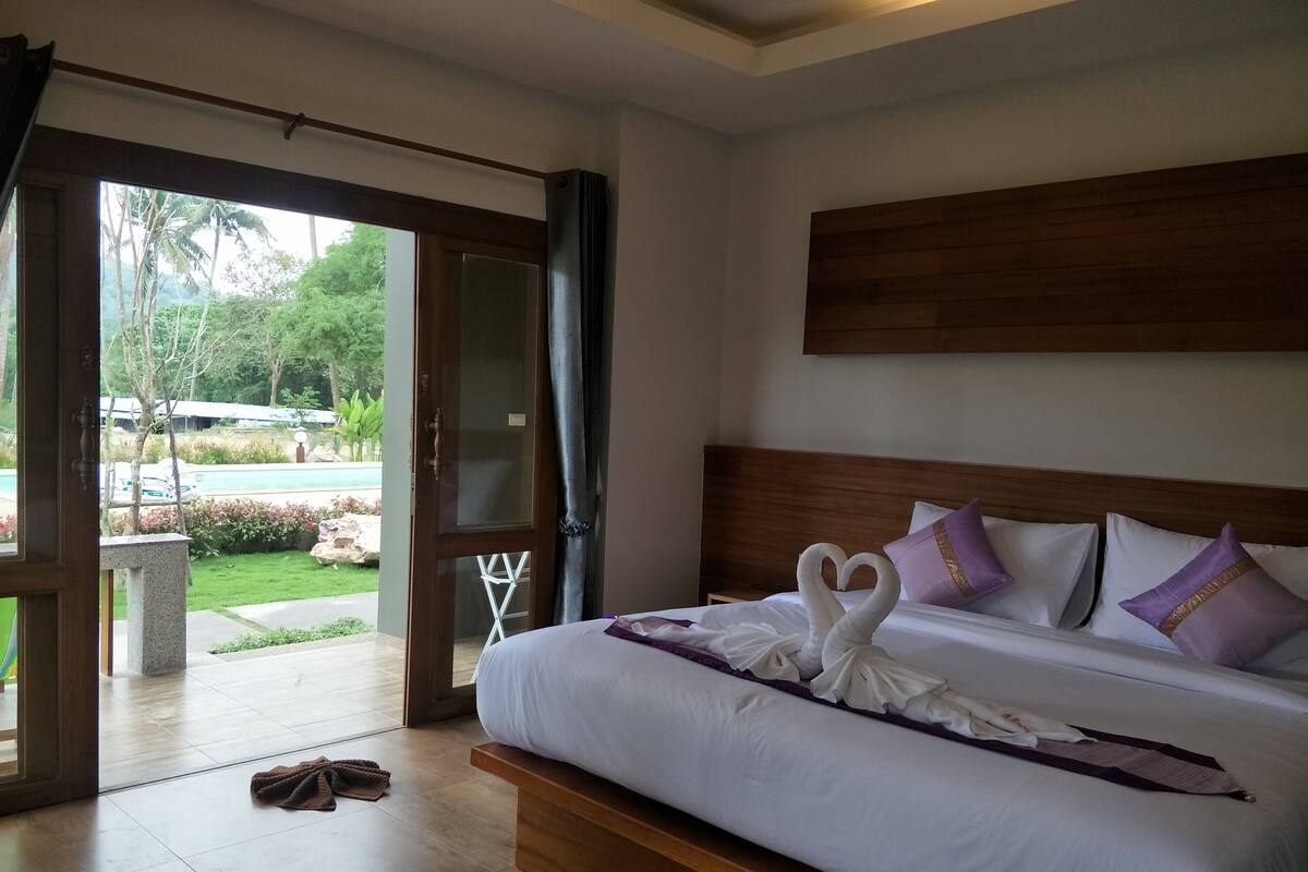 Une chambre d’hôtel avec un grand lit, une tête de lit en bois et une vue sur une piscine et la verdure environnante.