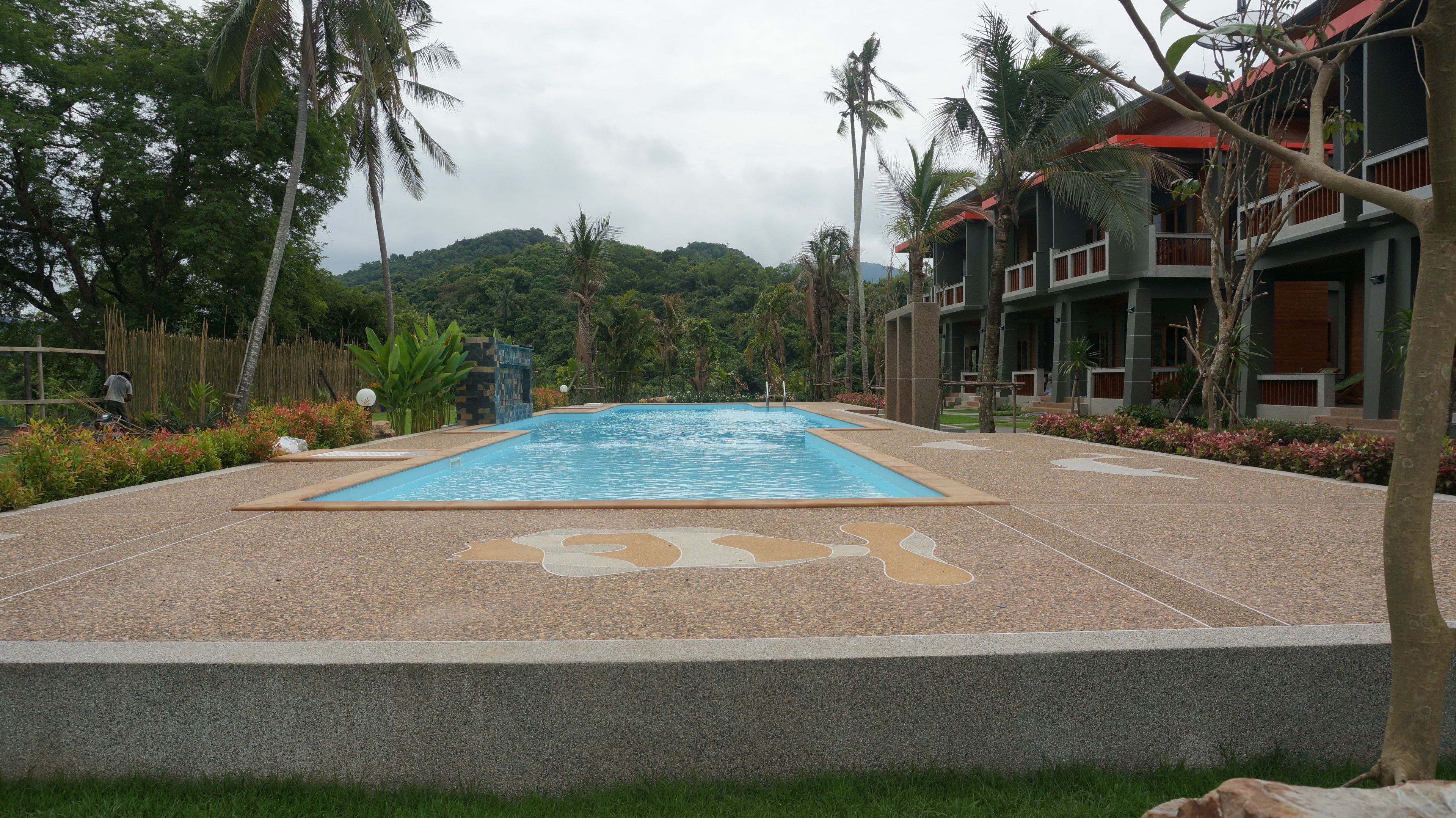 A swimming pool with a pebble border, surrounded by palm trees and greenery, with a view of a hilly landscape in the background.