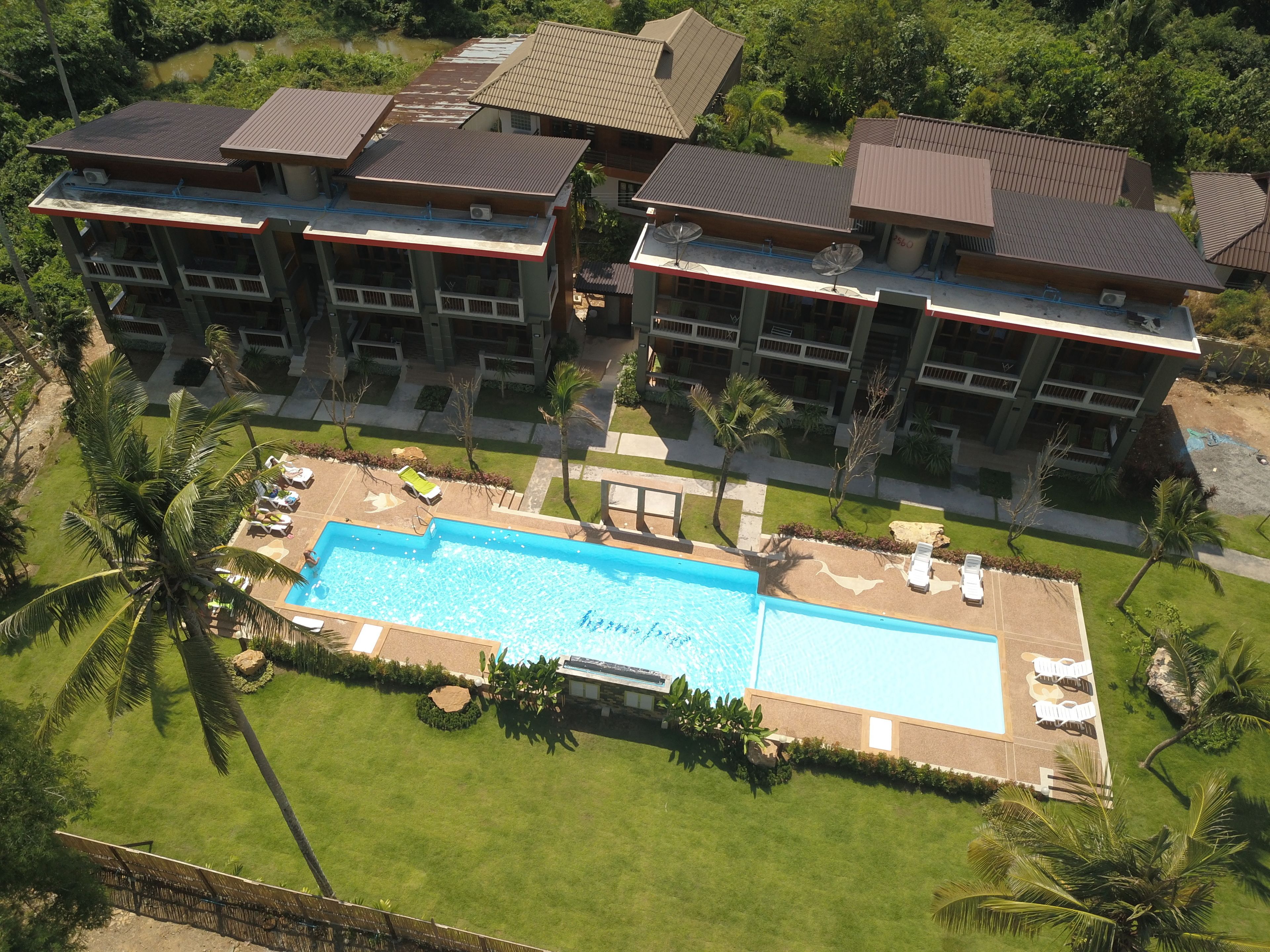 A large hotel with multiple buildings, a central swimming pool with sun loungers, and surrounding palm trees.