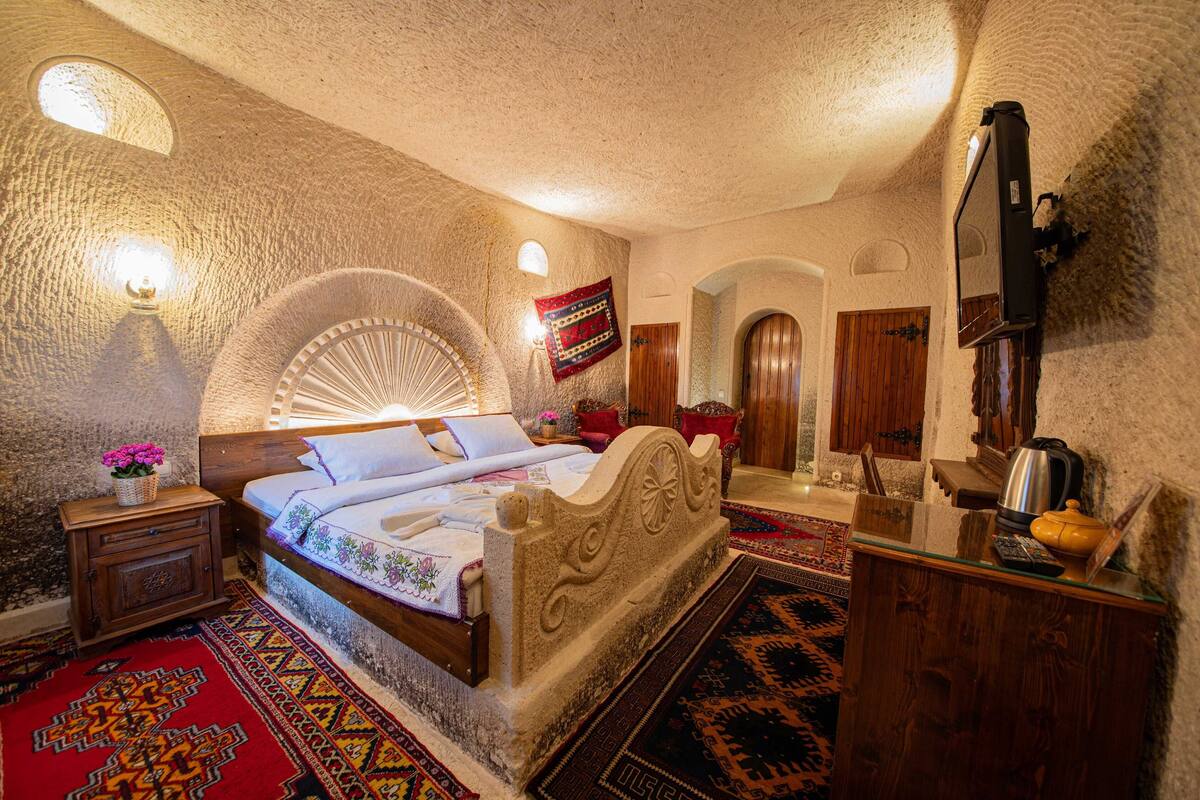 Quarto com cabeceira de pedra, cama com colcha estampada, mesa de cabeceira de madeira com um vaso de flores, tapete estampado em vermelho e preto, e uma porta de madeira ao fundo.