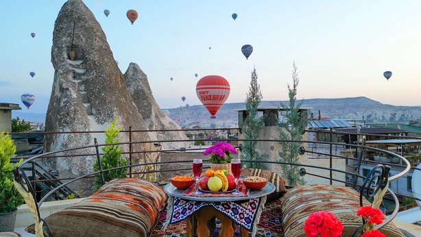 A terrace with a table set for dining, surrounded by traditional cushions and a view of rock formations and hot air balloons in the distance. - Safran Cave Hotel (Nevsehir)