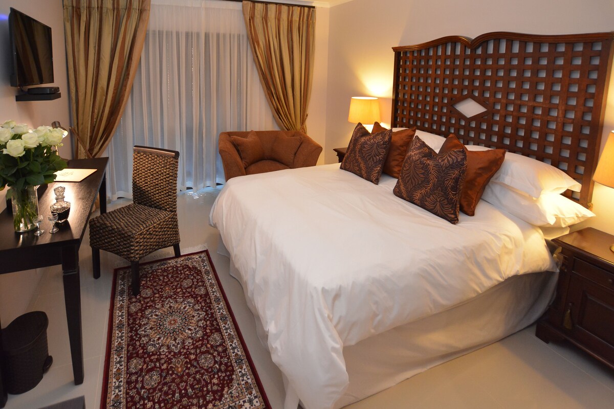A hotel room with a large bed, a wooden headboard, a desk with a lamp, a chair, and a decorative rug.