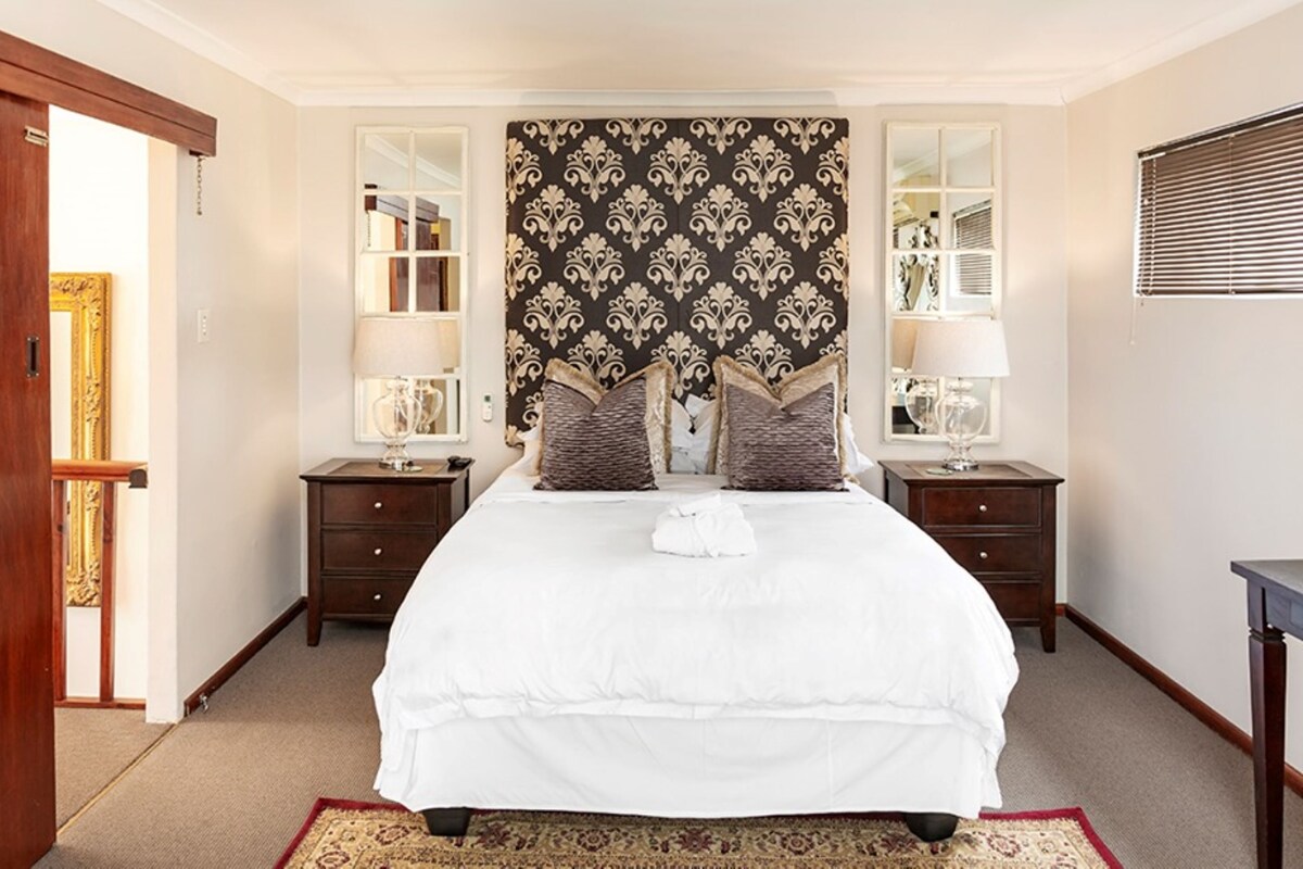 A neatly arranged bedroom with a large bed, two bedside tables with lamps, a decorative headboard, and a mirror on the wall.