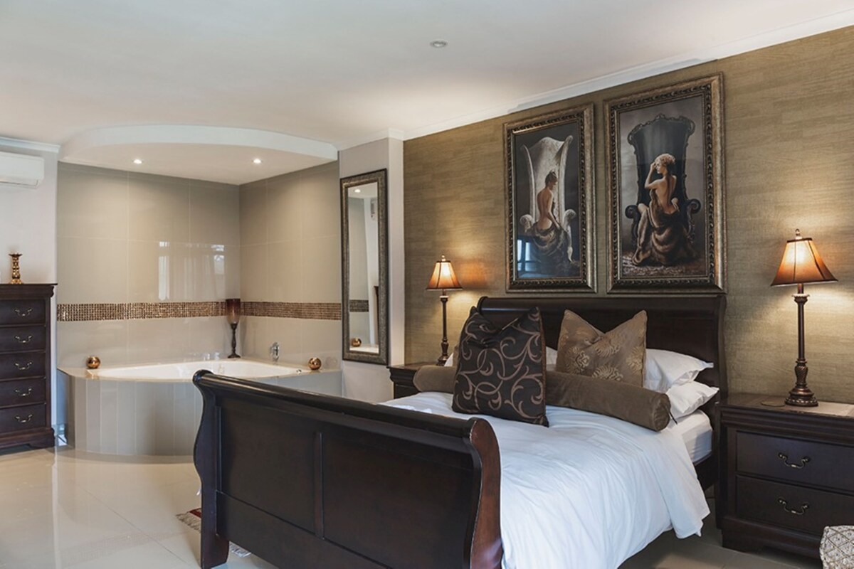 A bedroom with a large bed, two bedside tables, a nightstand, a mirror, and a wall-mounted artwork.