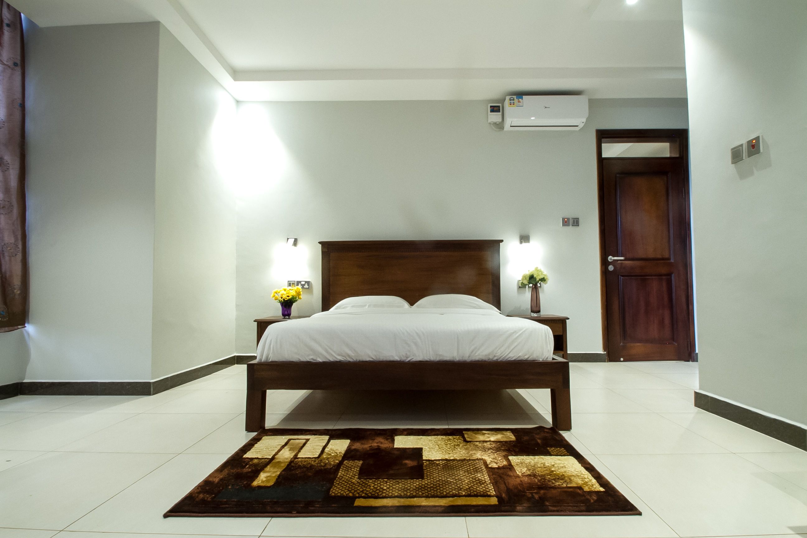 A bedroom with a large bed, a wooden headboard, a bedside table with a vase of flowers, and a wall-mounted air conditioner.