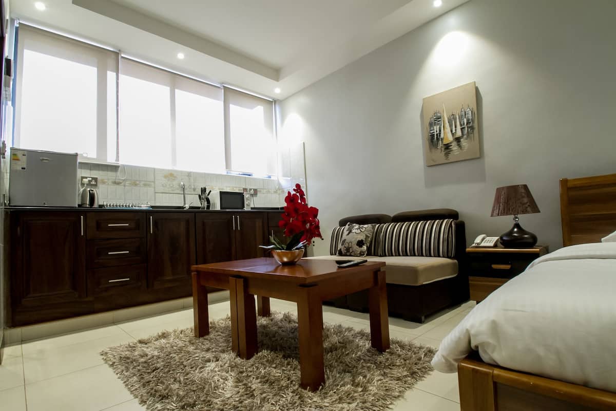 A modern kitchen with dark cabinets, a wooden table with a vase of red flowers, a sofa, and a bed in a bedroom.