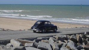 A vintage car parked on a beach with a rocky foreground and ocean in the background. - Le Tertre (La Feuillie)