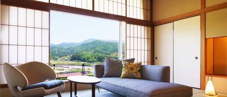 A room with tatami flooring, a sofa, a chair, and a table. A large window offers a view of a green landscape.