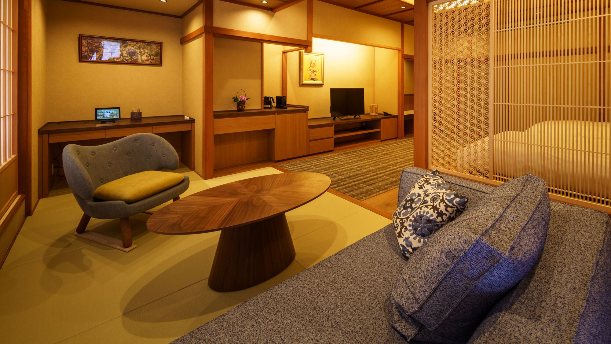 Una habitación tradicional japonesa con suelo de tatami, una mesa de madera baja, un sofá con cojines estampados, un televisor y un biombo decorativo.