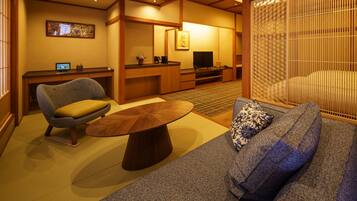 Una habitación tradicional japonesa con suelo de tatami, una mesa de madera baja, un sofá con cojines estampados, un televisor y un biombo decorativo.
