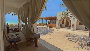 A beachside seating area with a view of the ocean, wooden furniture, and a covered outdoor space. - Coco Beach Hotel (Jambiani)