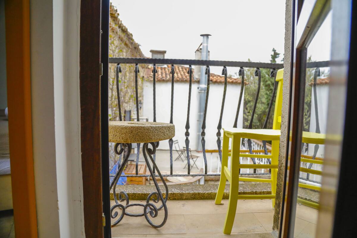 A balcony with a round table and a yellow chair, a metal railing, and a view of buildings with red-tiled roofs.