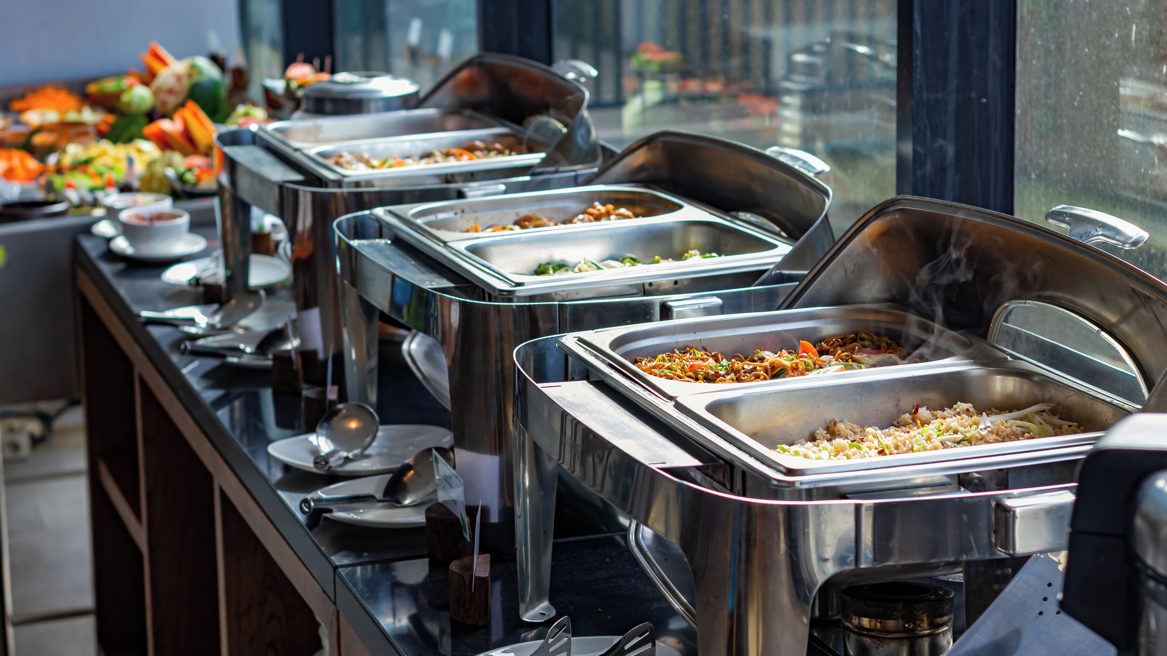 A buffet setup with multiple stainless steel chafing dishes containing various foods, arranged on a counter with a window view in the background.