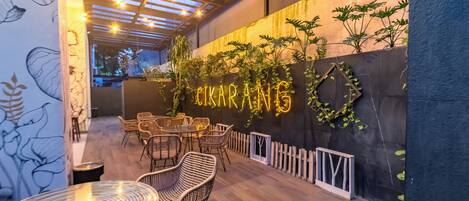 Un espacio exterior con asientos de mimbre y mesas, una pared con vegetación y letreros de neón, además de una pérgola decorada con luces colgantes.