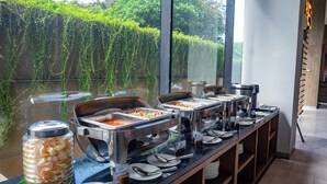 Disposição de buffet com diversos recipientes de comida e utensílios, um pote de lanches e vista para o verde através de grandes janelas.