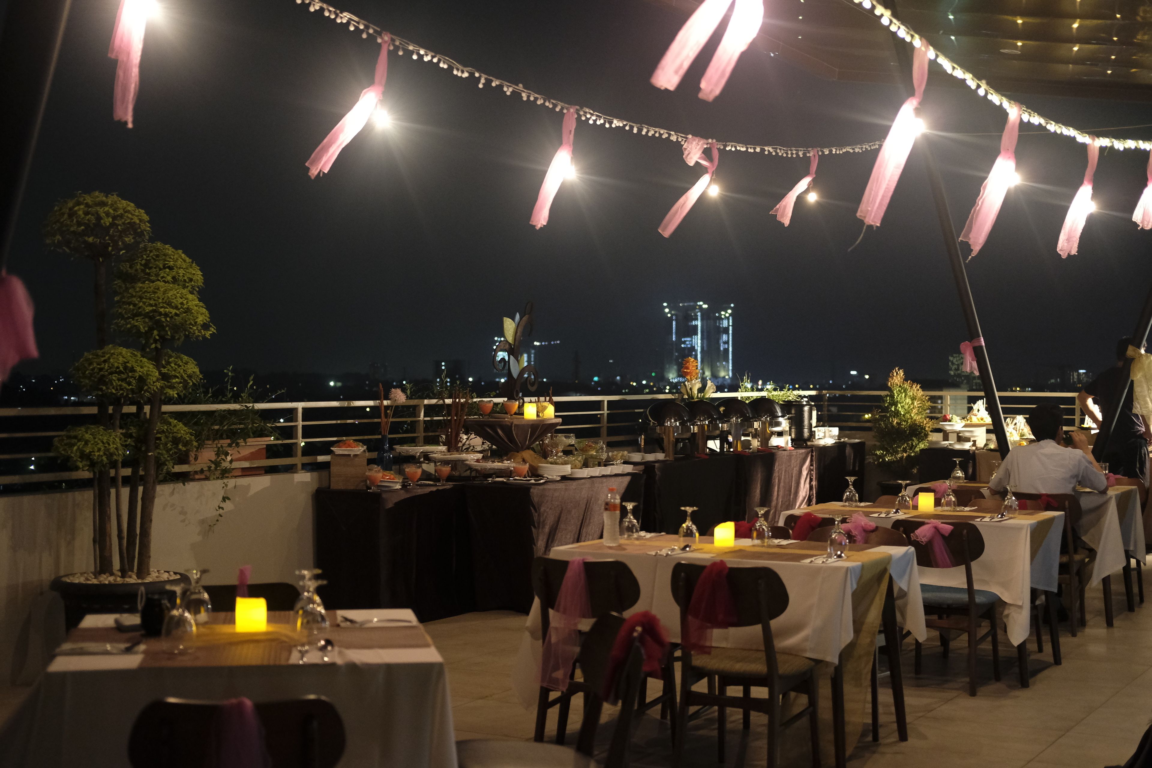 An outdoor dining area with tables set for two, string lights overhead, and a view of the city skyline in the background.