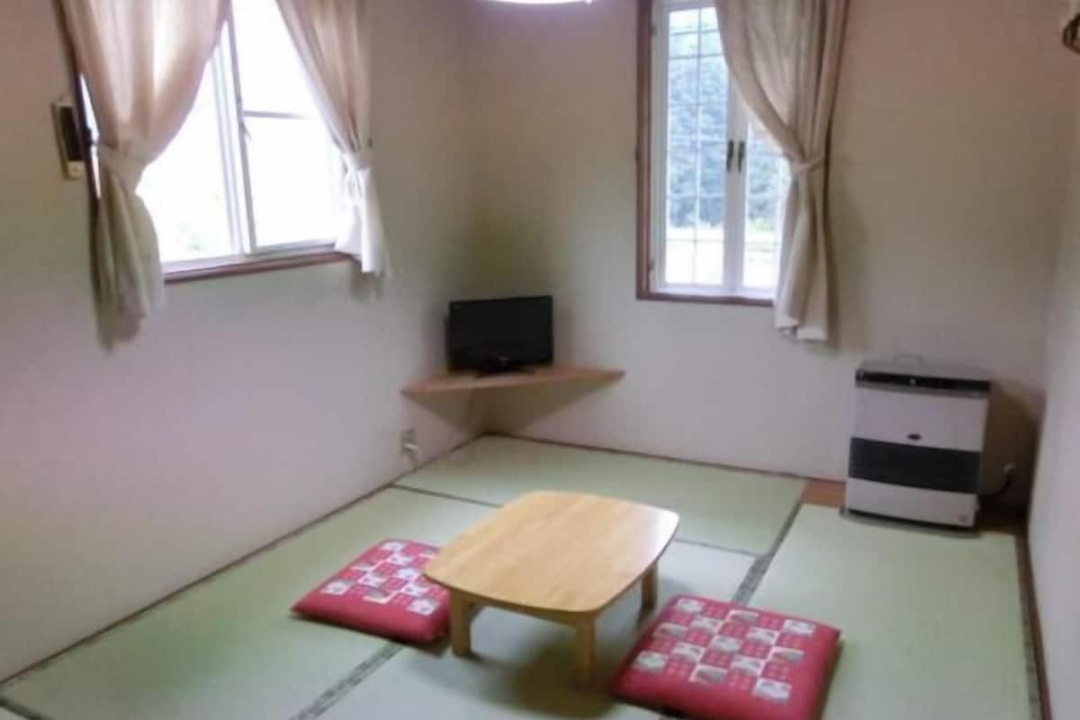 Una habitación con suelo de esteras de tatami, una mesa de madera, dos cojines rojos, un televisor sobre una estantería y un pequeño electrodoméstico blanco.