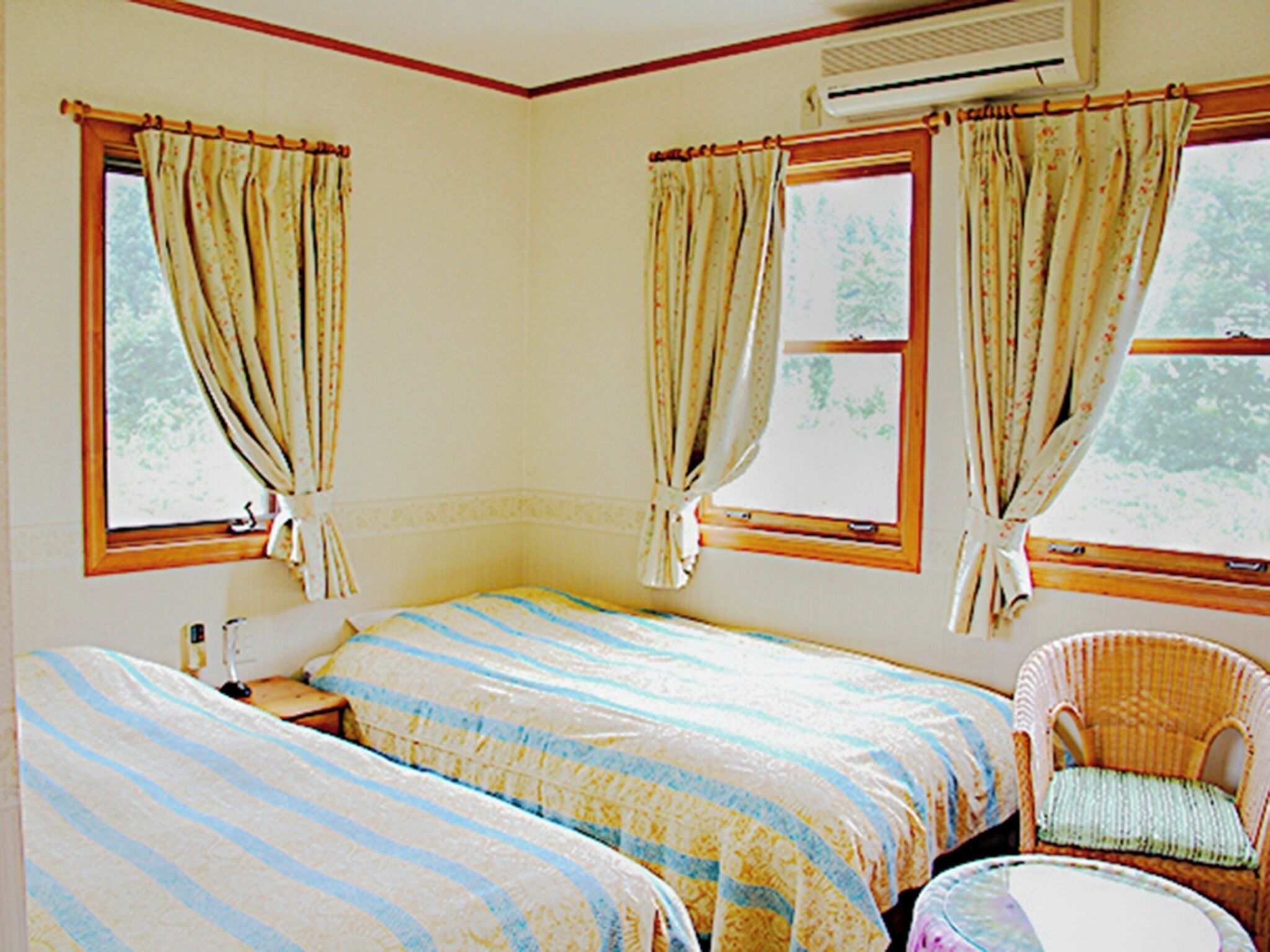 Quarto com duas camas, uma cadeira de vime, uma mesa pequena e um aparelho de ar condicionado.