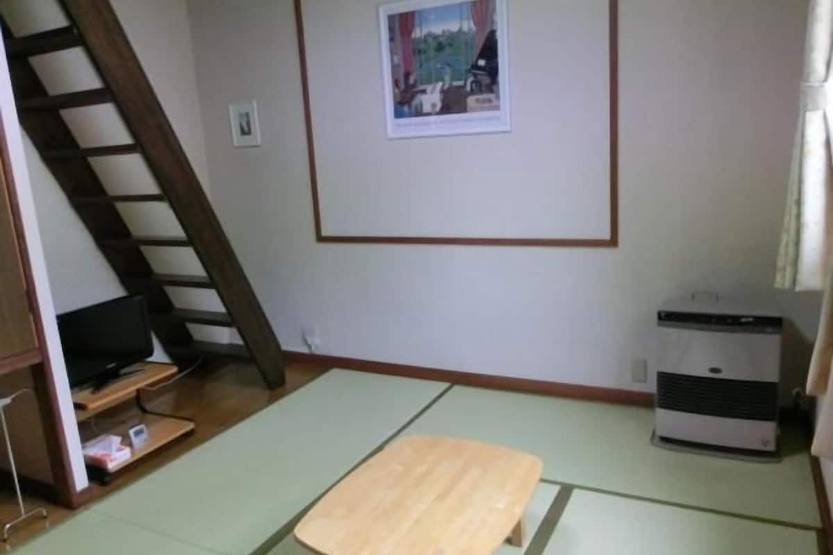 Una habitación con esteras de tatami, una mesa de madera, un televisor sobre un pedestal, un calentador y un cuadro enmarcado en la pared.