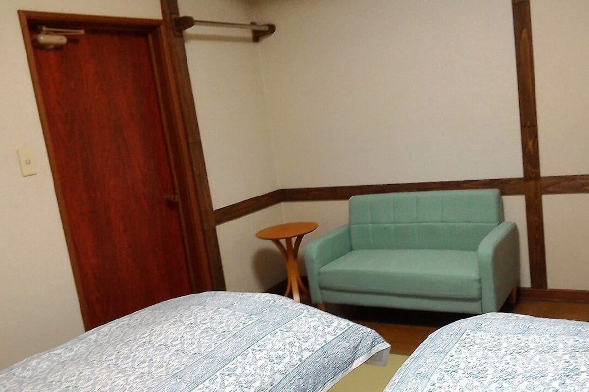 Una habitación pequeña con dos camas, una puerta de madera, una mesita redonda y un sillón color turquesa.
