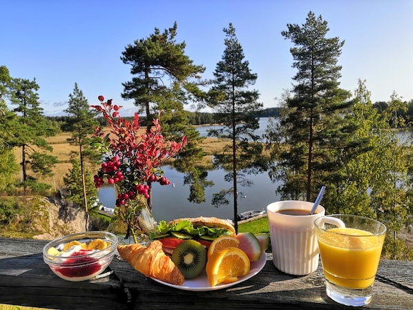 Un montaje para picnic con un croissant, fruta, un vaso de zumo de naranja y una taza de café sobre una mesa de madera con vistas a un lago rodeado de árboles.