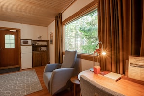 Una habitación acogedora con techo de madera, una ventana con cortinas, un escritorio con lámpara, una silla, una mesita y un refrigerador.