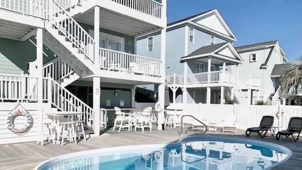 Un área de piscina con terraza de madera, mobiliario exterior blanco y un edificio de dos pisos con balcones.